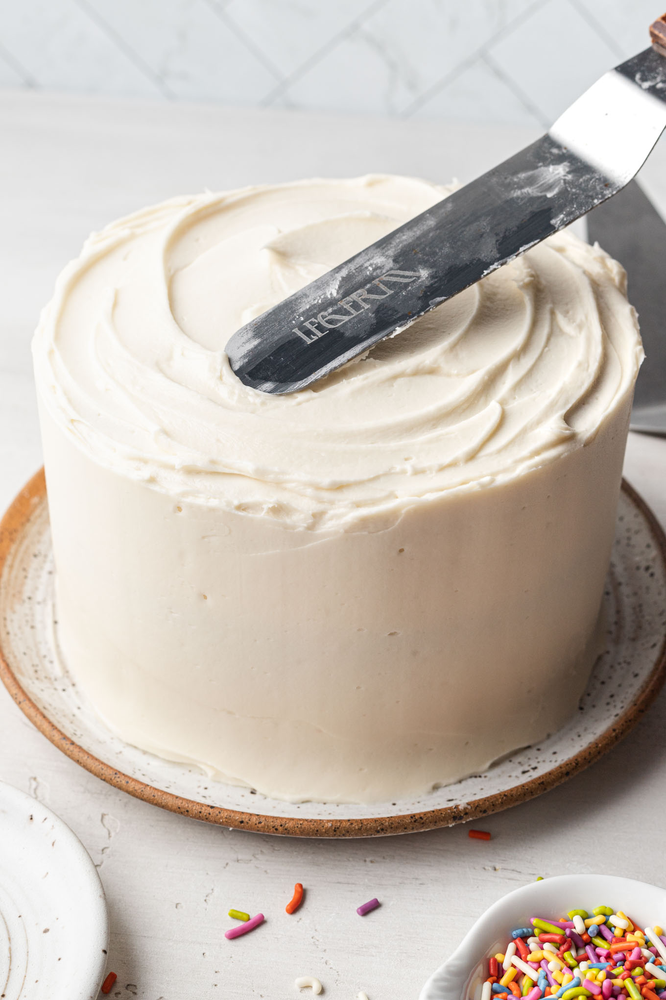 A mini vanilla cake on a speckled white plate being frosted.