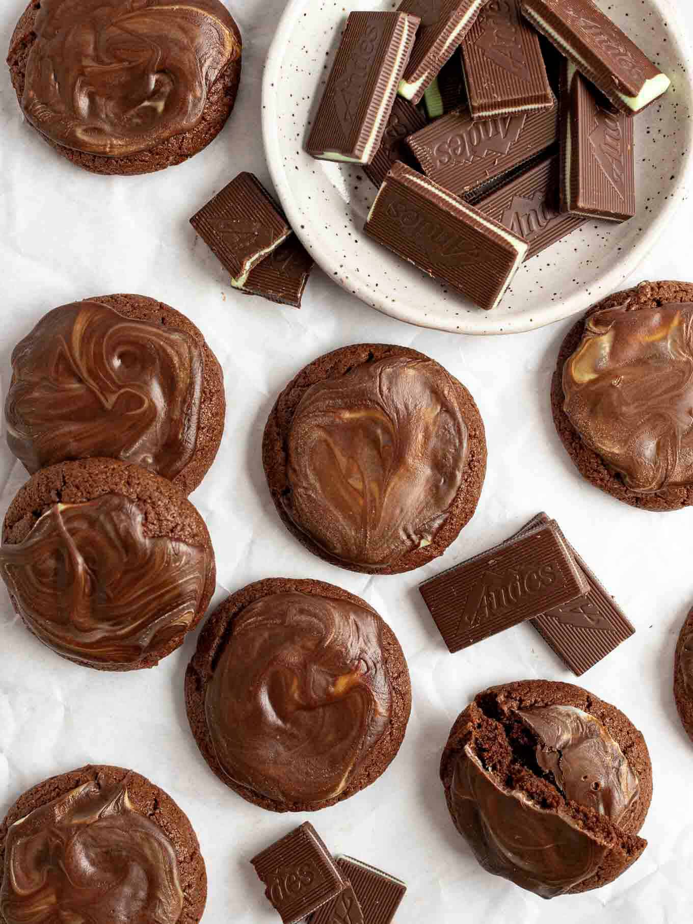 An overhead view of several Andes mint cookies spread out on a piece of parchment paper. A bowl of Andes mints is sitting next to the cookies.