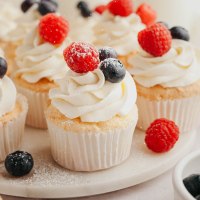 Several angel food cupcakes topped with whipped cream and fresh berries on a marble serving platter.