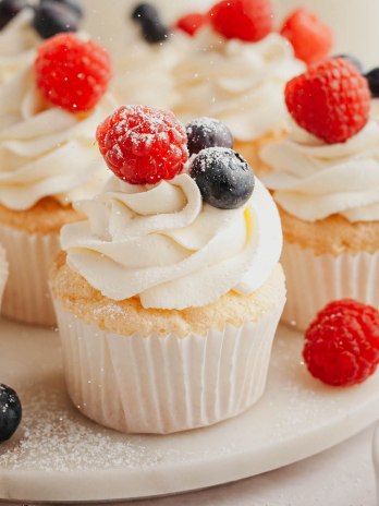 Several angel food cupcakes topped with whipped cream and fresh berries on a marble serving platter.