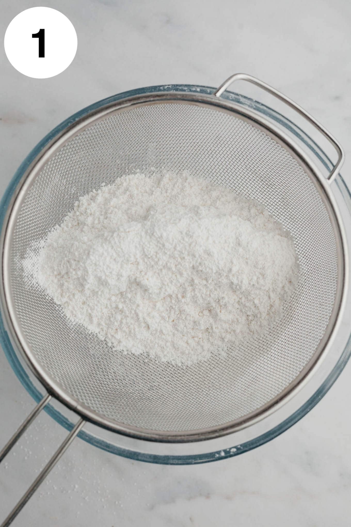 An overhead view of flour and powdered sugar in a fine mesh sieve over a mixing bowl. 