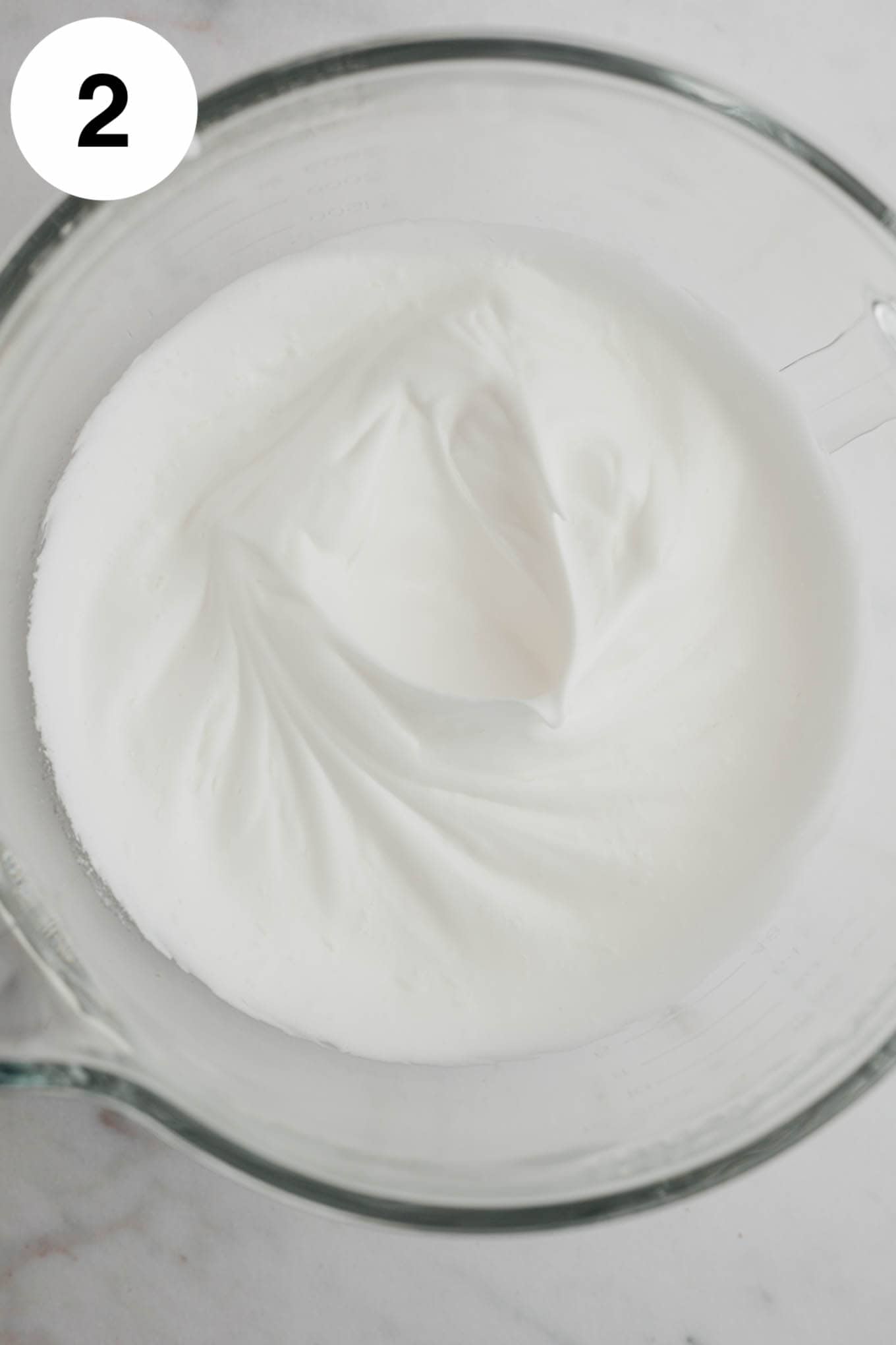 An overhead view of whipped egg whites in a glass mixing bowl. 