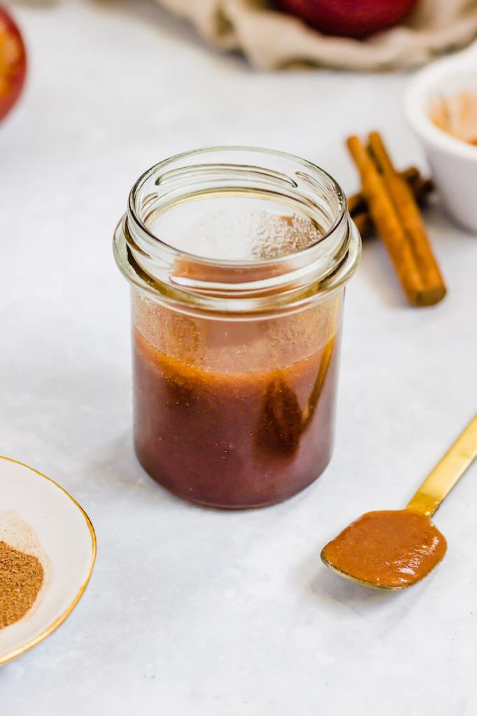 A jar filled with apple butter. A spoon with apple butter and cinnamon sticks are around the jar.