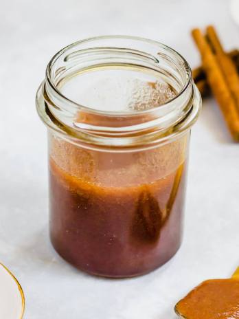 A jar filled with apple butter. A spoon with apple butter and cinnamon sticks are around the jar.