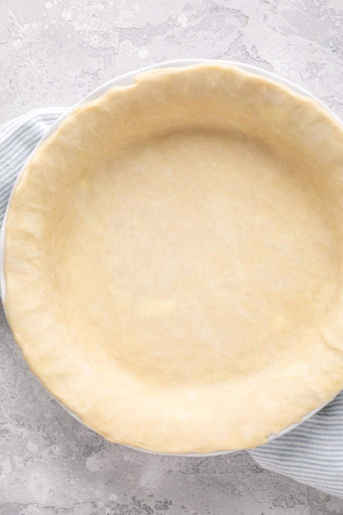 An unbaked pie crust inside of a white pie dish. The dish is placed on top of a blue and white striped napkin.