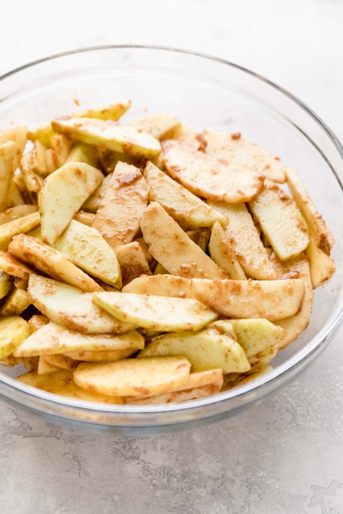 Apple slices coated in a mixture of flour, spices, and lemon juice in a glass bowl.