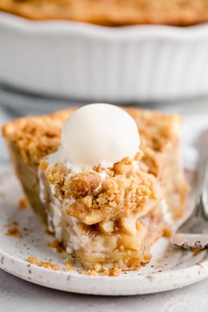 A slice of apple crumble pie topped with a scoop of ice cream that's melting off the sides. The pie has a bite taken out of it to show the inside of the pie.