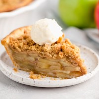 A slice of apple crumble pie topped with a scoop of ice cream on a white plate. Two apples and the rest of the pie are in the background.