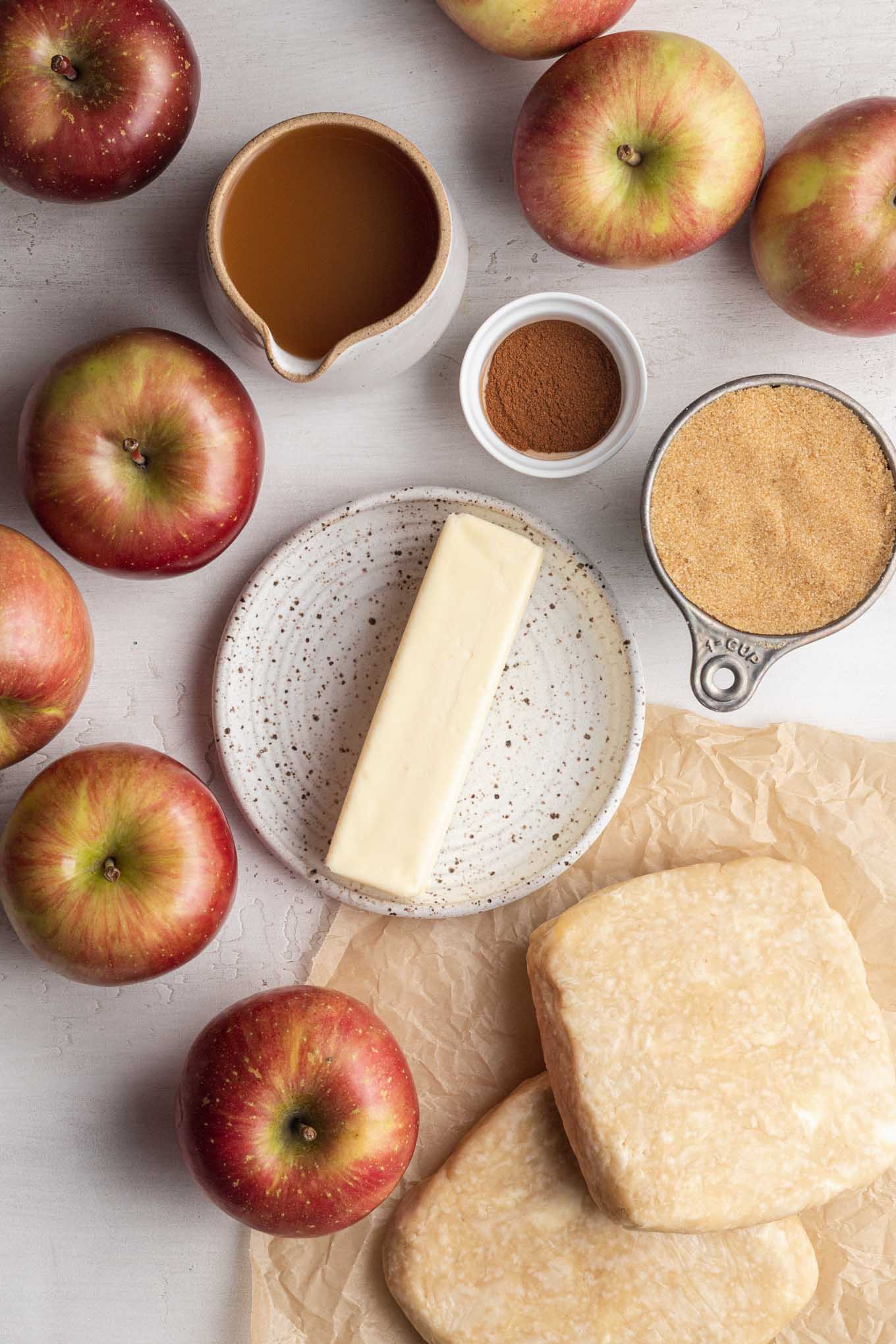 An overhead view of apples, apple cider, brown sugar, cinnamon, butter, and pie crust.