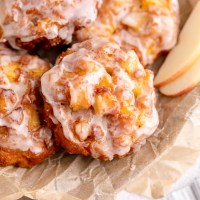 Several glazed apple fritters on top of a piece of brown parchment paper in a metal dish. A couple of apple slices are resting next to the fritters.