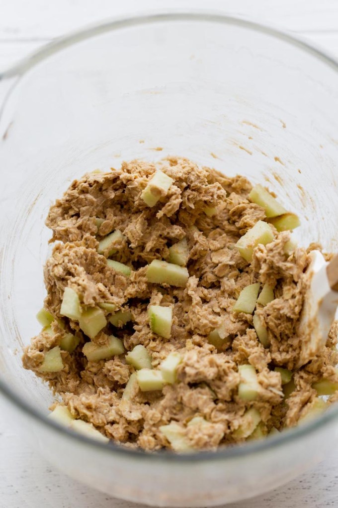 A glass mixing bowl holding cookie dough with chopped apples folded into it.