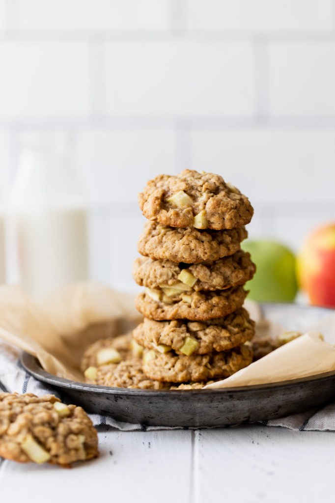 A stack of apple oatmeal cookies resting in a metal baking dish. A container of milk and a couple of apples rest in the background.