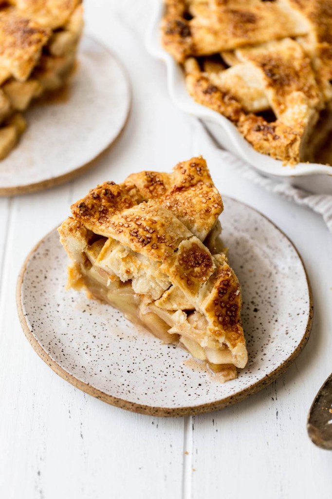 A single slice of apple pie on a plate with the rest of the pie in a white dish behind it.