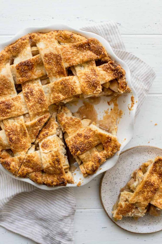 A baked apple pie in a white pie dish with one slice removed and put on a plate.