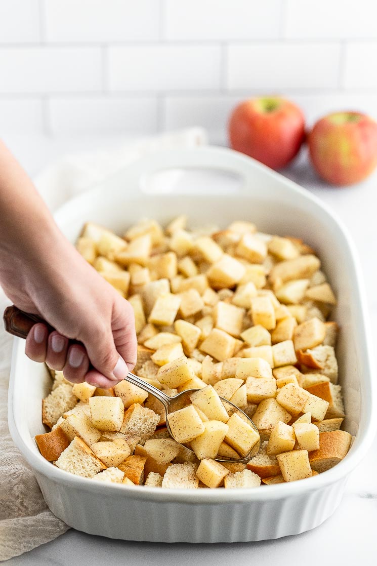 Chopped apples being adding on top of cubed bread in a baking pan.