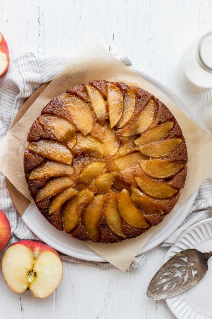 An overhead view of an apple upside-down cake with sliced apples and milk around it.