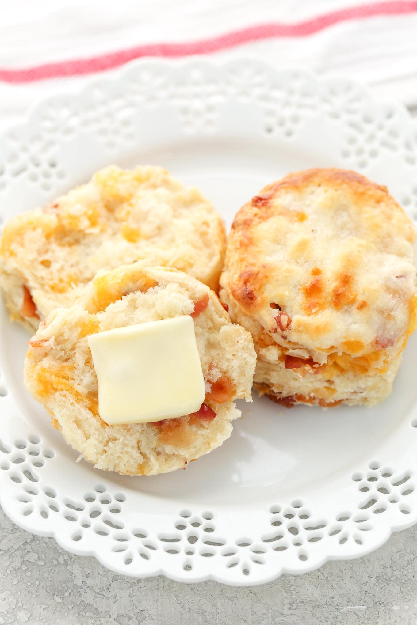 Two bacon cheddar biscuits on a white plate. One biscuit is cut in half and has a pat of butter on it. 