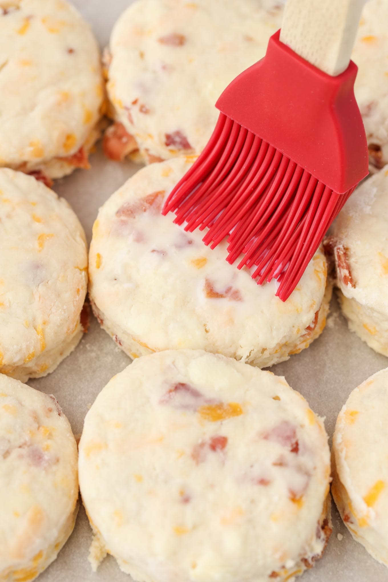 Three rows of unbaked cheddar cheese biscuits being brushed with buttermilk. 