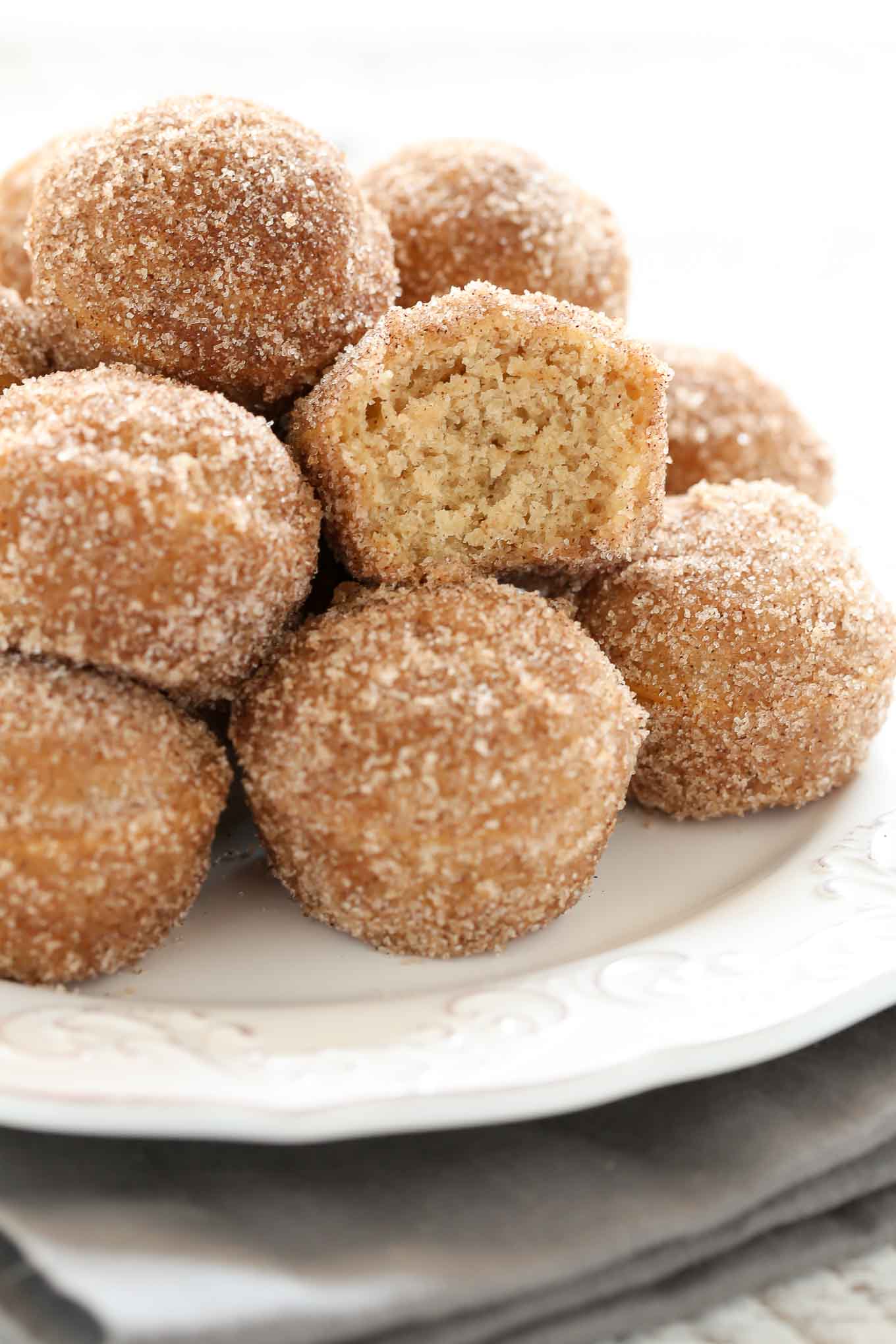 Close up shot of pile of cider donut holes on a white plate. The middle donut hole has a bite missing. 