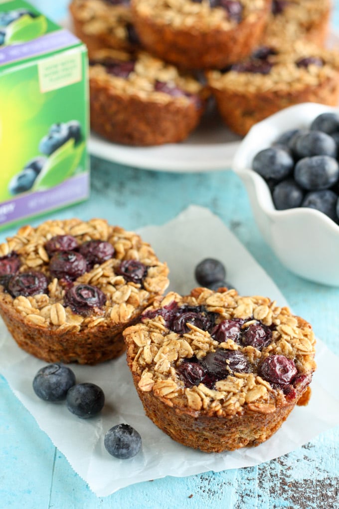 Two baked oatmeal cups topped with blueberries on a piece of parchment paper. More oatmeal cups, a bowl of berries, and a box of tea rest in the background. 