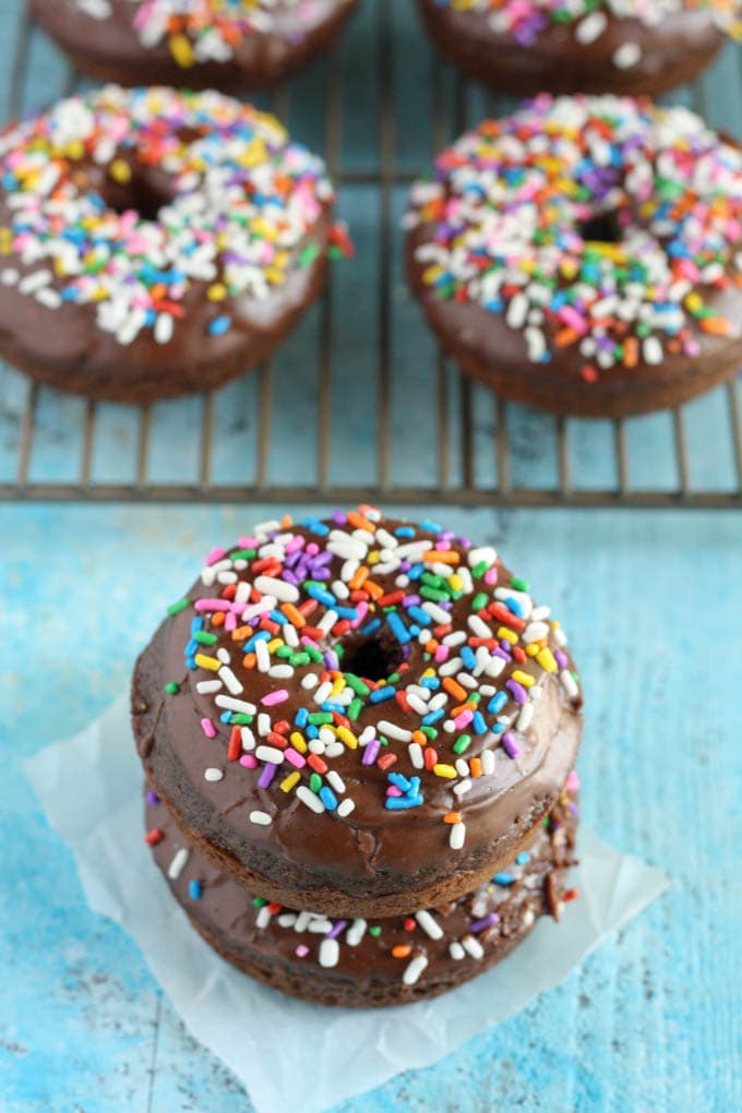 A stack of two frosted chocolate donuts on a square of parchment paper. More donuts are on a cooling rack in the background. 