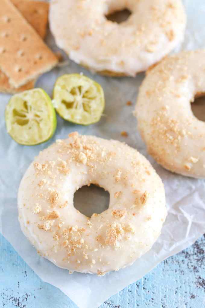 Three key lime donuts on parchment paper. Lime halves and graham crackers are in the background. 