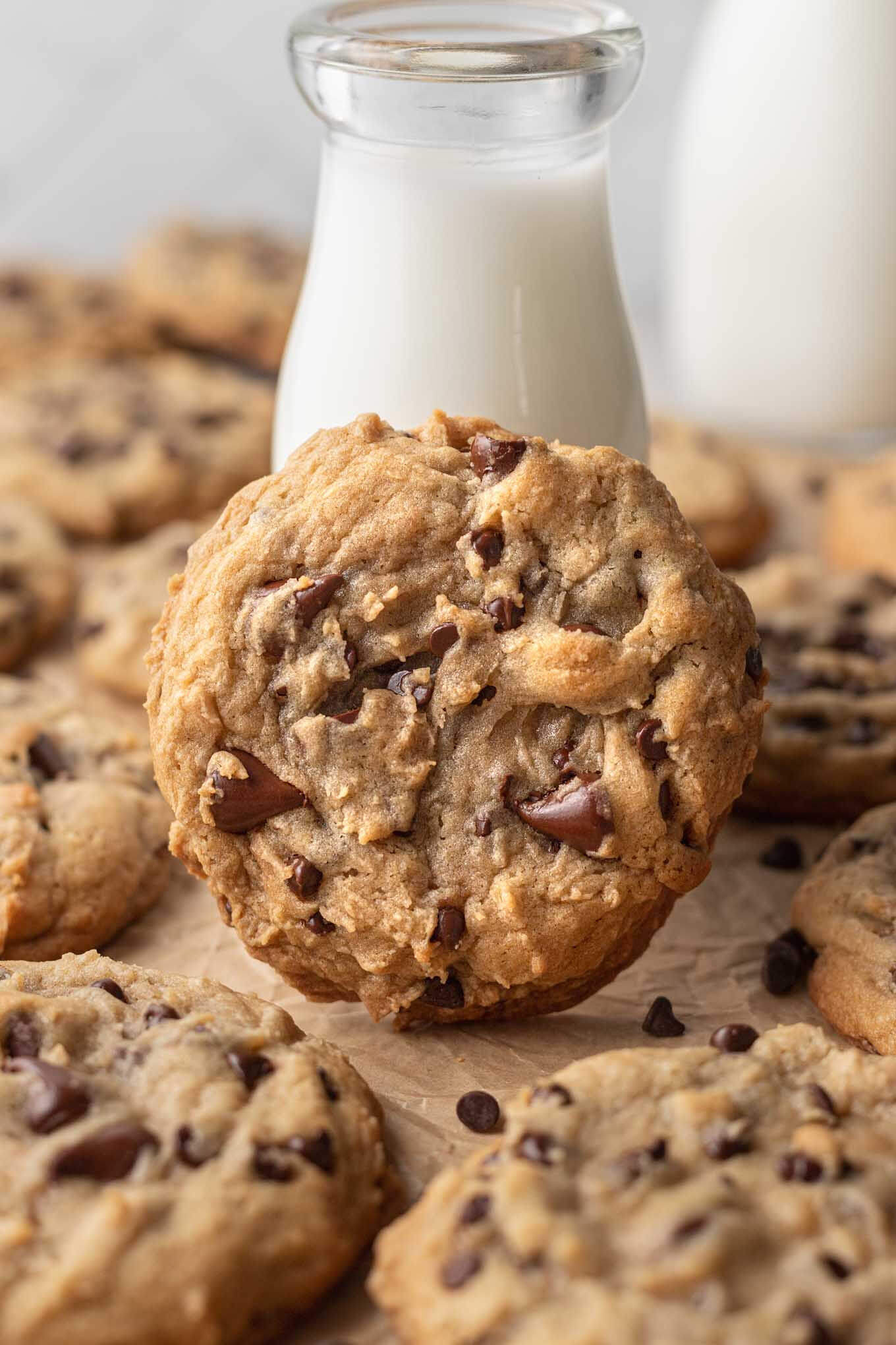 A large bakery style chocolate chip cookie leaning against a tall glass of milk. More cookies are surrounding it.