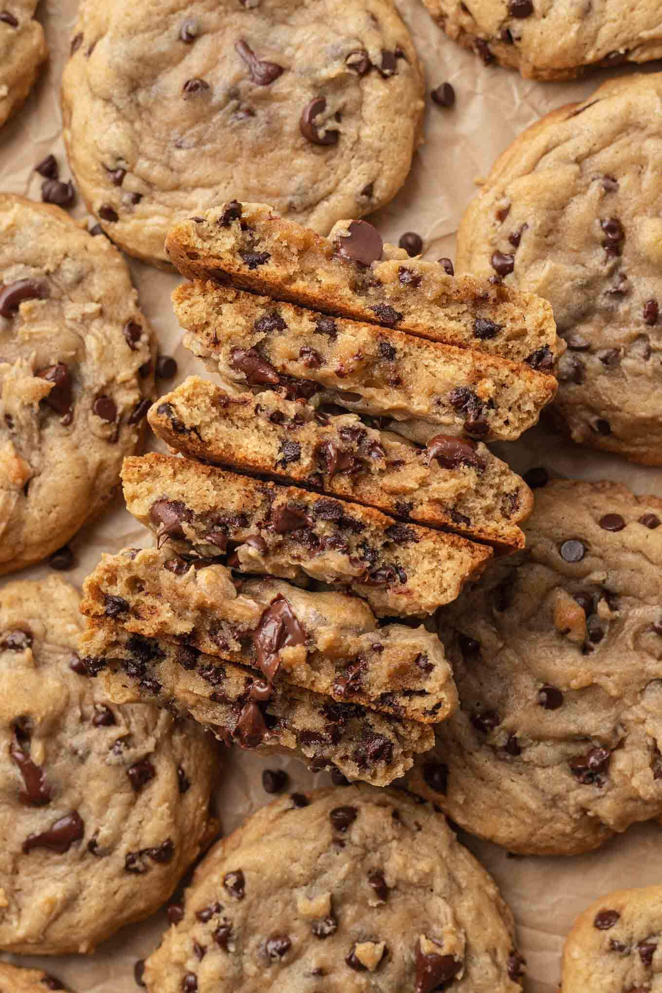 An overhead view of several chocolate chip cookies on a piece of brown parchment paper. Three of the cookies are broken in half to show the texture.