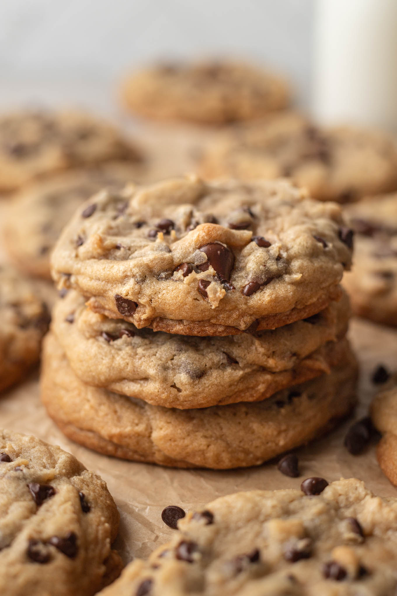 A stack of three bakery style chocolate chips cookies on a piece of brown parchment paper. More cookies are surrounding the stack of cookies.