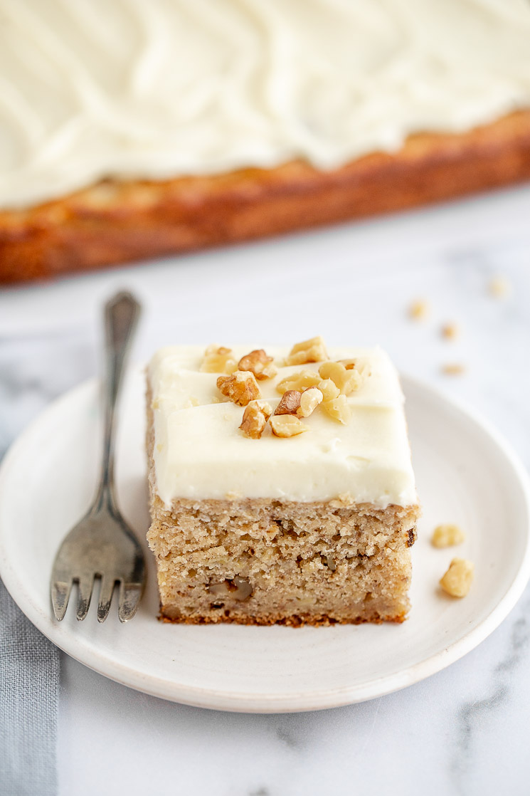 A piece of banana cake sitting on an antique white plate and the rest of the cake in the background on a marble surface.