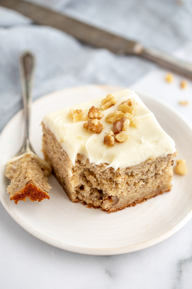 A square piece of banana cake topped with cream cheese frosting and extra walnuts that has a bite sized piece taken out on an antique fork.