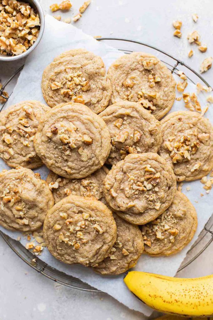 An overhead view of a pile of banana nut cookies on a wire cooling rack. Chopped walnuts are scattered around the cookies. A banana rests on the side. 