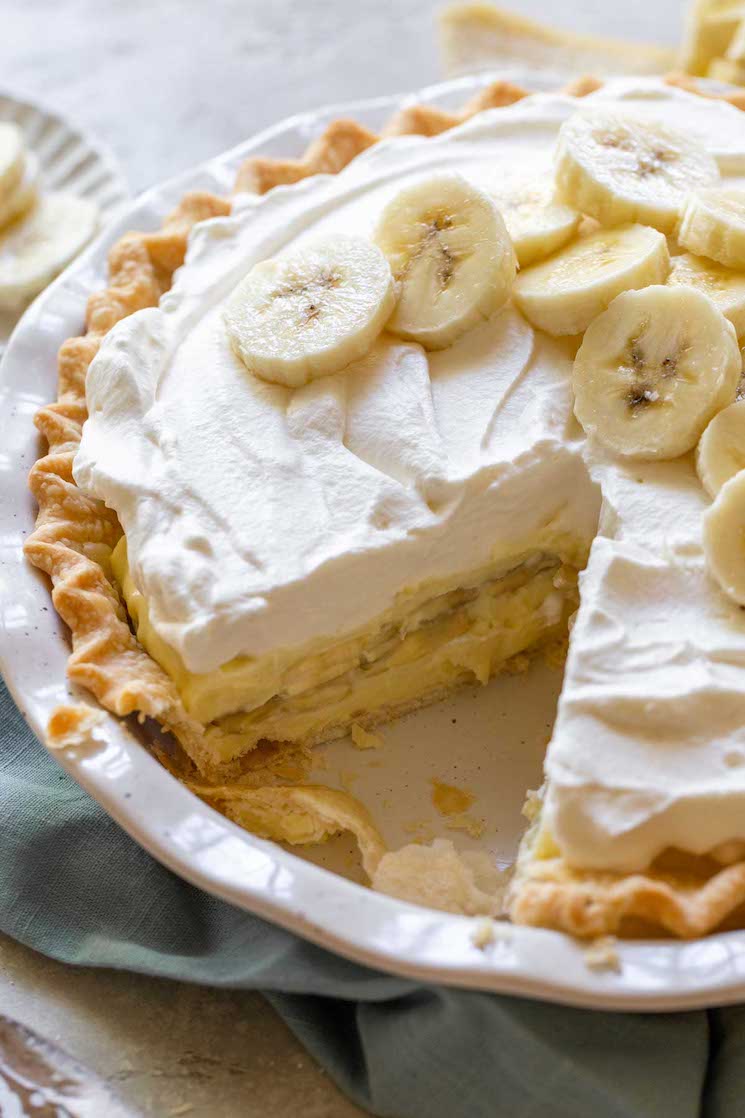 A finished banana cream pie with one slice cut out to show the layers of filling, bananas, and homemade whipped cream.