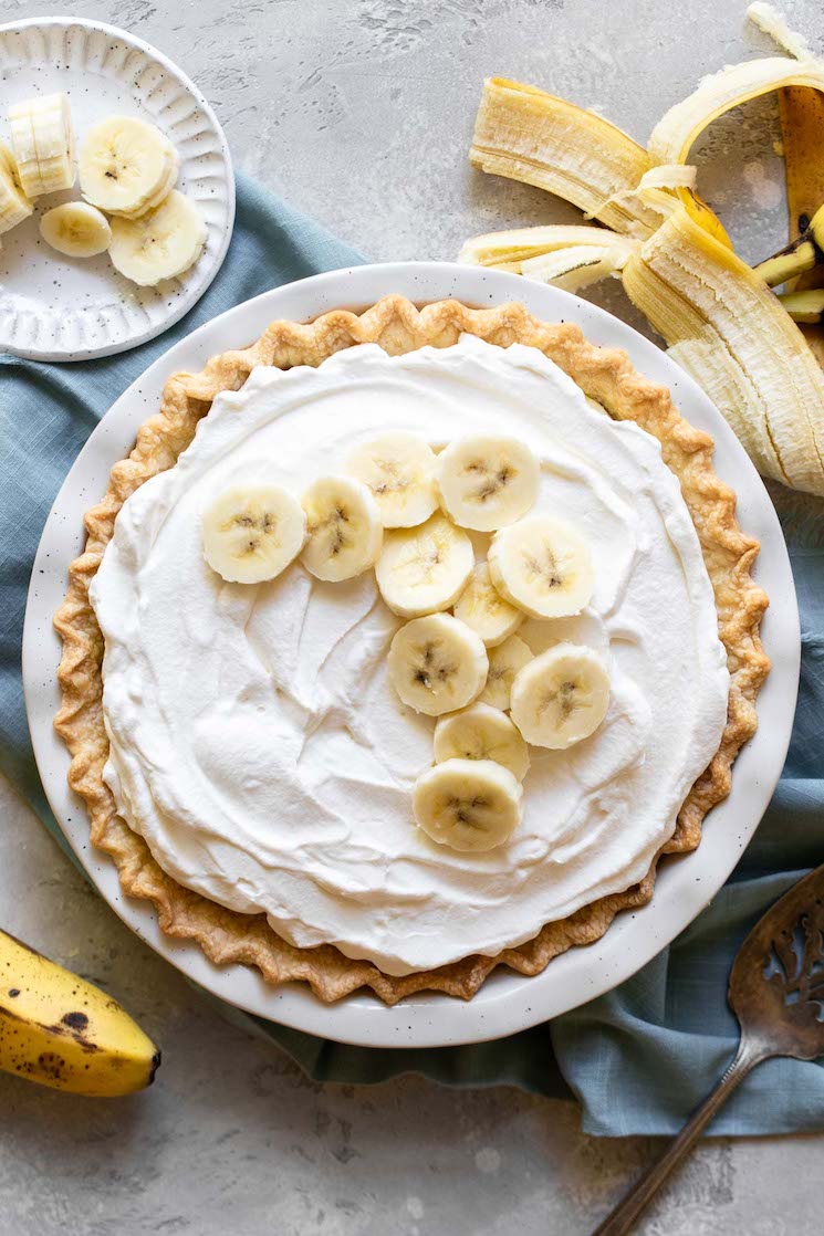 A finished banana cream pie in a white pie plate with banana slices on top with more bananas and a pie server next to it.