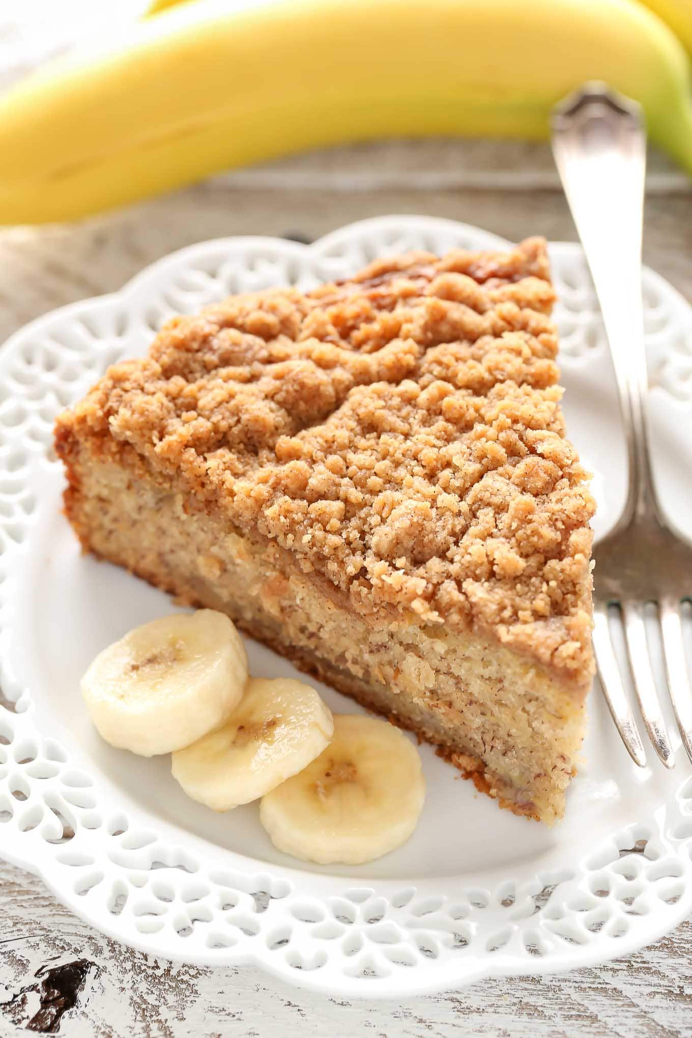 A slice of banana crumb cake, a fork, and three banana slices on a white plate. A banana rests in the background. 