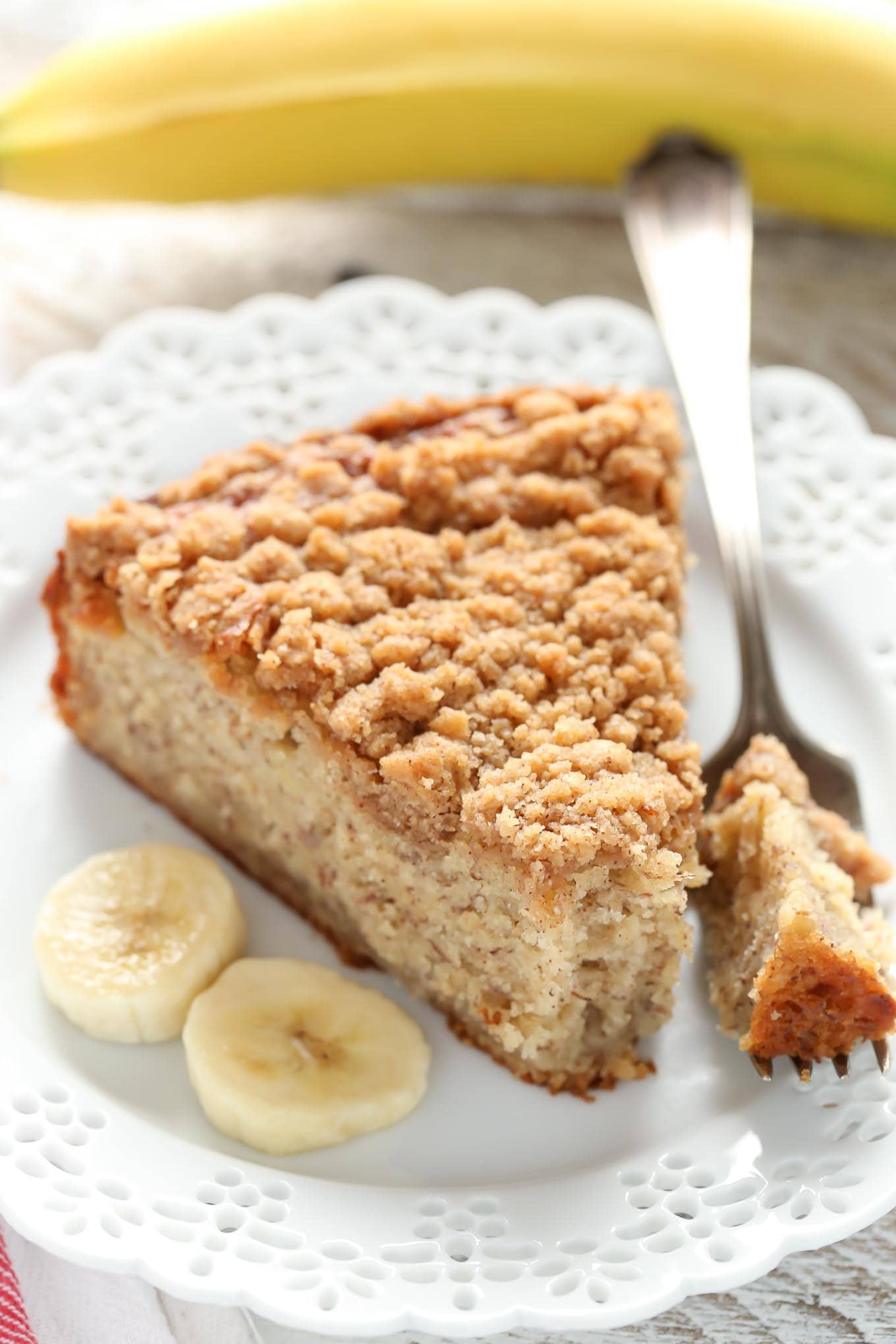 A slice of banana crumb cake, a fork, and three banana slices on a white plate. A banana rests in the background. 