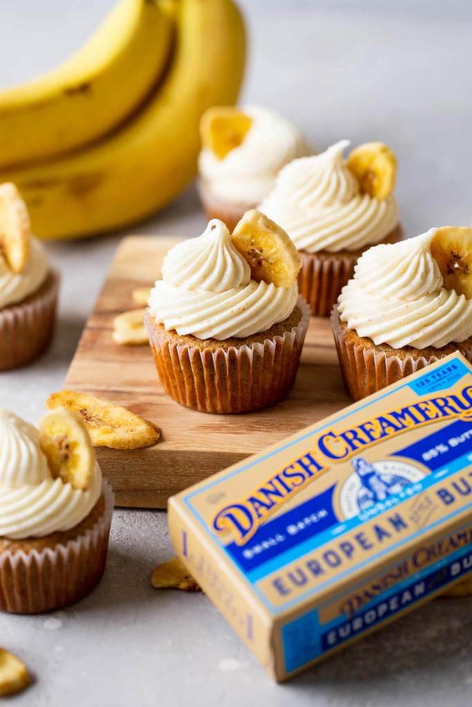 Side view of banana cupcakes with cream cheese frosting on a wooden board. A bunch of bananas rest in the background. 