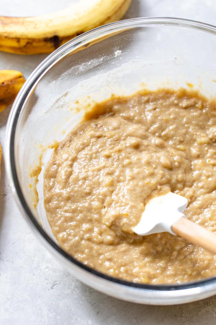 The dry and wet ingredients being mixed together in a glass bowl with an extra banana in the background.