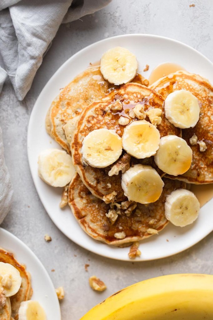 Overhead view of fluffy banana pancakes topped with banana slices and chopped walnuts on a white plate. 