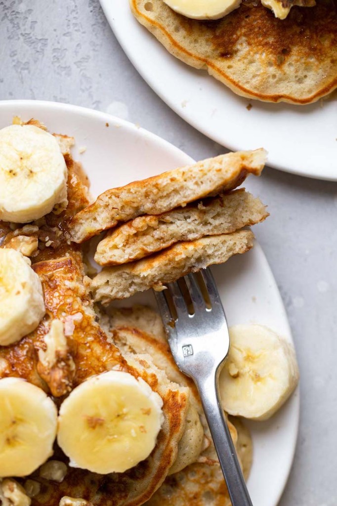 Overhead view of fluffy banana pancakes on a white plate. A fork has speared a bite of pancake. 