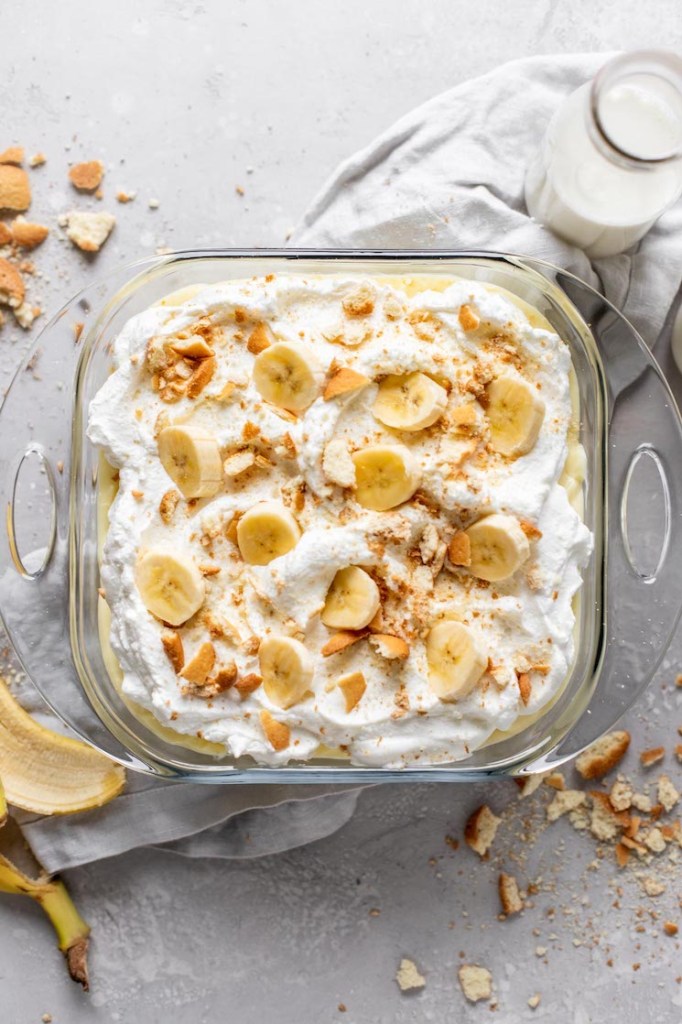 Overhead view of a baking dish filled with banana pudding from scratch. Crushed Nilla Wafers and a banana peel surround the baking dish. 