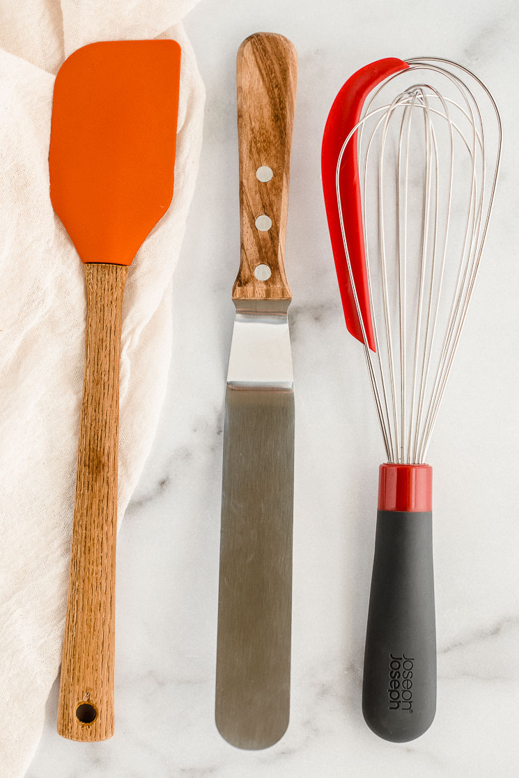 A rubber spatula, an icing spatula, and a whisk resting on top of a marble counter.