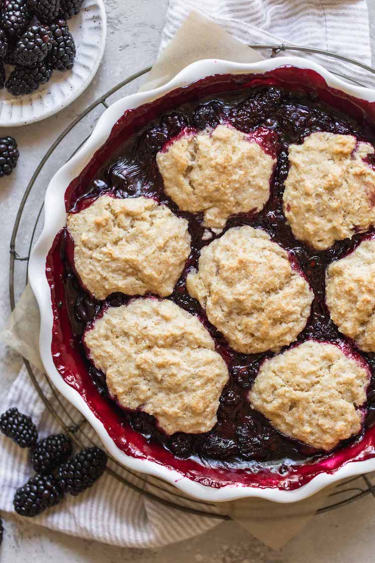 A finished blackberry cobbler in a round baking dish with extra blackberries around it.