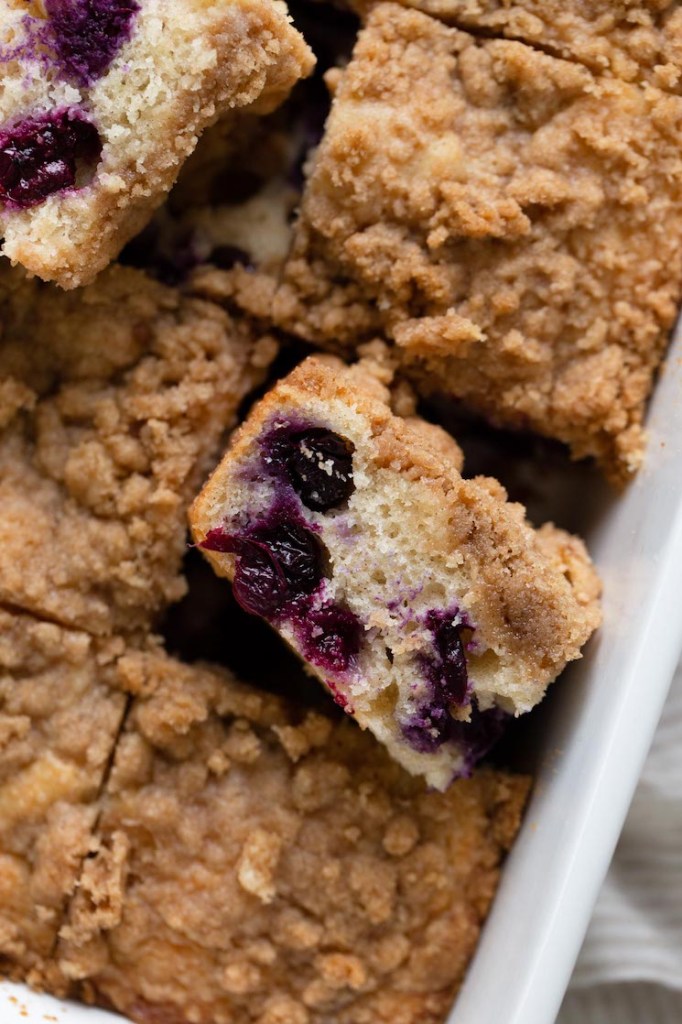 An overhead view of a blueberry streusel coffee cake in a white baking dish. The cake has been sliced and some slices rest on their sides. 