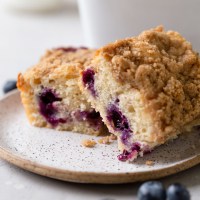 A side view of two slices of blueberry coffee cake on a speckled plate. Fresh blueberries rest in the foreground.