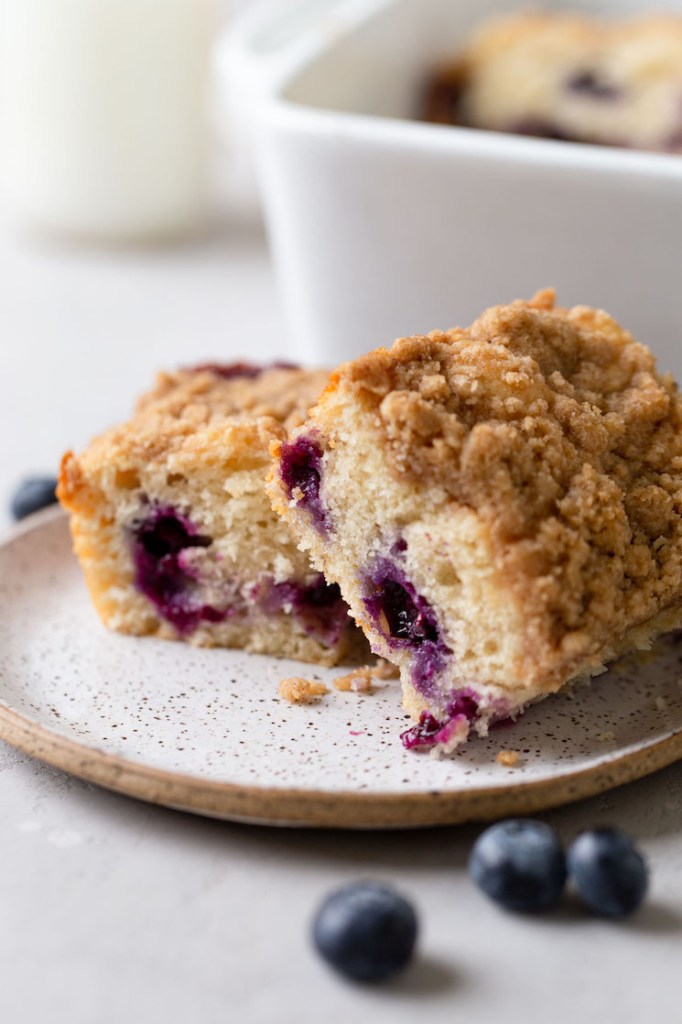 A side view of two slices of blueberry coffee cake on a speckled plate. Fresh blueberries rest in the foreground and a pan of cake rests in the background. 