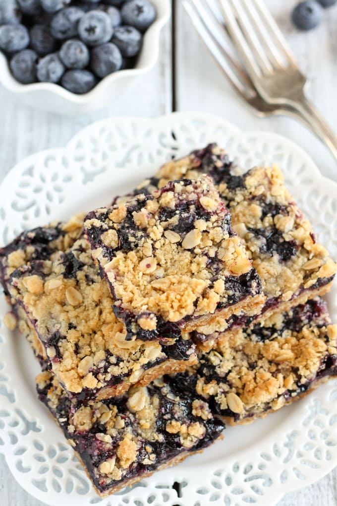 Overhead view of a stack of blueberry bars on a white plate next to a bowl of fresh berries and two forks. 