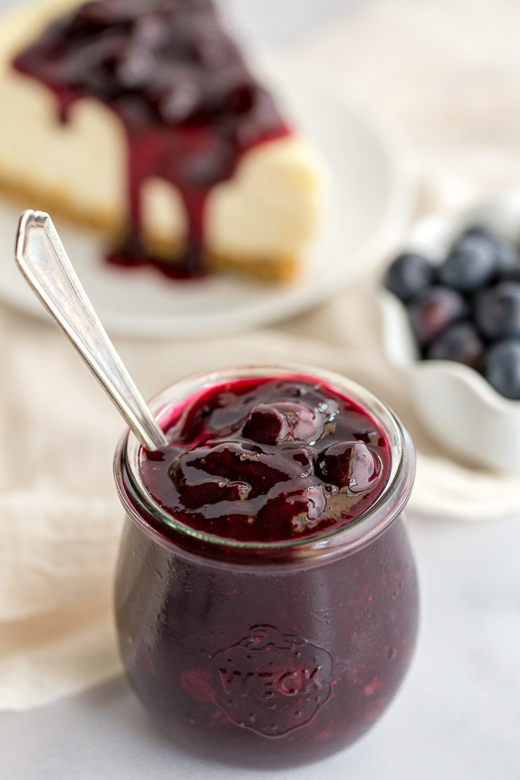 A jar of homemade blueberry sauce with more blueberries and a slice of cheesecake in the background.