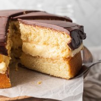 A sliced Boston cream pie on a cake stand. One slice is being removed.