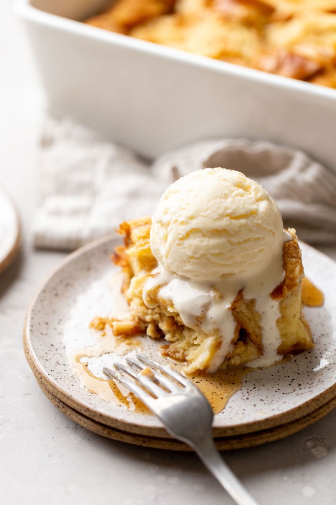 A slice of bread pudding topped with vanilla ice cream. A bite has been taken out of it to show the texture.